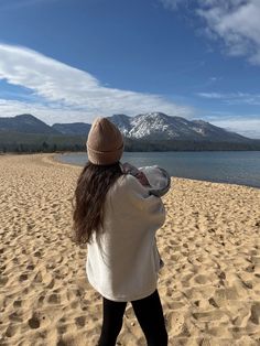 a woman standing on top of a sandy beach next to the ocean with mountains in the background