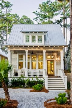 a small white house with a porch and stairs leading up to the front door, surrounded by pine trees