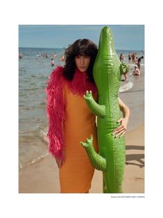 a woman in an orange dress is holding a large green alligator statue on the beach