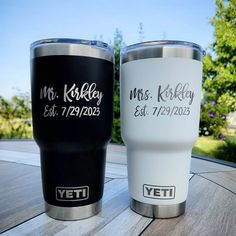 two personalized yeti tumblers sitting on top of a wooden table in front of trees