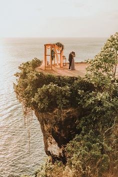 two people standing on the edge of a cliff overlooking the ocean with greenery around them