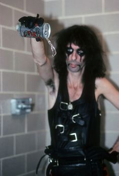 a man with black hair and makeup is holding a can in his hand while standing next to a brick wall