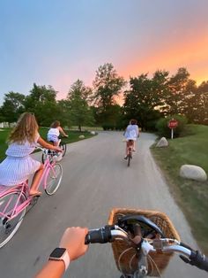 three people riding bikes down a road with grass and trees in the background at sunset