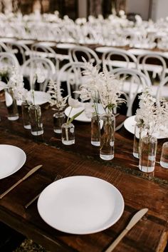 the table is set with white flowers in vases and plates on it, along with silverware