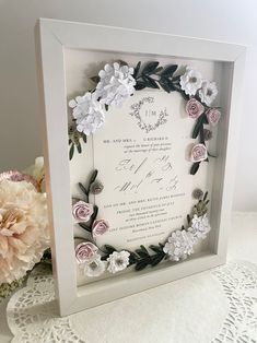 a white frame with flowers and greenery on the inside is shown next to a pink flower bouquet