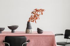 a table topped with a vase filled with red flowers next to two black chairs and a pink table cloth