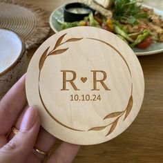 a person holding up a wooden coaster with the initials r and f on it in front of a plate of food