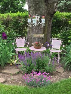 two chairs and a table in the middle of a garden with purple flowers around it