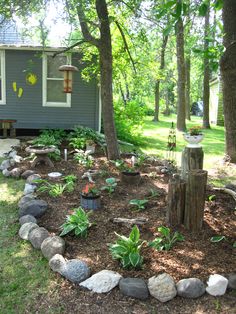 a garden with rocks and trees in front of a blue house on a sunny day