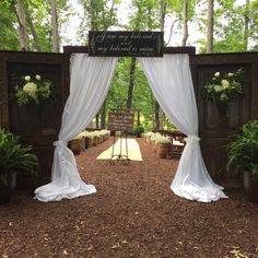 an outdoor ceremony with white drapes and greenery on the side, surrounded by trees