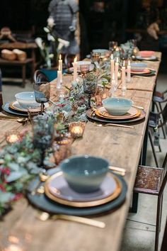 a long wooden table topped with plates and place settings covered in greenery next to candles