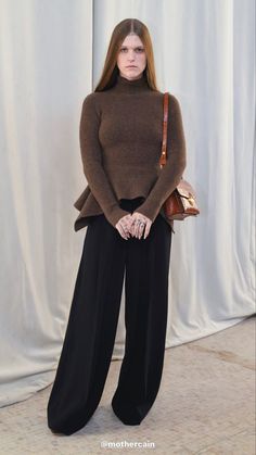 a woman standing in front of a white curtain wearing black pants and a brown sweater