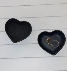 two heart shaped trays with rings in them on a white wooden background, one is black and the other is gold