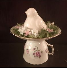 a white bird figurine sitting on top of a bowl filled with flowers and greenery