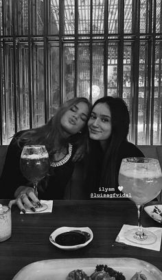 two women sitting at a table with food and drinks in front of them, posing for the camera