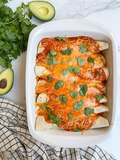 an enchilada casserole in a white dish with avocado and cilantro on the side