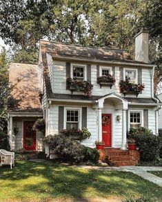 a white house with red front door and window boxes on the windowsill is surrounded by greenery