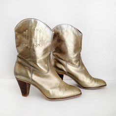 Vintage 70s metallic gold slip on cowboy boots with stacked wooden heels. The interior is lightly padded. Made of man made materials. Made in Spain. No marked size but estimated to fit a size 8.5. Please use the measurements below to ensure an accurate fit. Measurements: approximately 10" long (insole), 3" at the ball, and 2" at the heel of the foot. Heel is about 3" high. Condition: There is some color fade at the opening and back of the heels, giving the boots an ombre effect. There are also s Gold Heeled Boots With Round Toe For Fall, Gold Round Toe Heeled Boots For Fall, Vintage Party Boots, Gold Snip Toe Boots For Fall, White And Gold Cowboy Boots, Gold Western Boots With Almond Toe, Western Style Gold Heeled Boots, Western Gold Boots With Pointed Toe, Luxury Gold Women's Cowboy Boots