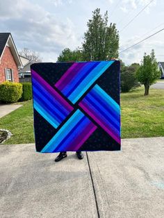 a person holding up a colorful quilt in front of a house with grass and trees