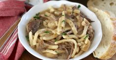 a white bowl filled with pasta and meat on top of a wooden table next to bread