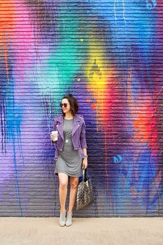 a woman standing in front of a colorful wall holding a coffee cup and looking at the camera