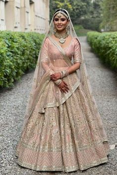 a woman in a pink lehenga standing on a gravel road with bushes behind her