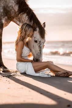 a woman is sitting on the beach next to a horse and kissing it's face