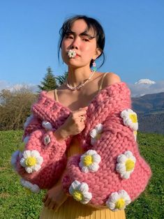 a woman standing in the grass wearing a pink sweater with white flowers on it and holding her hands to her chest