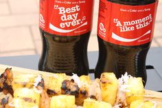 two bottles of soda sitting on top of a wooden table next to some grilled food