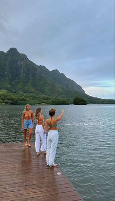 three women are standing on a dock pointing at the water with mountains in the background