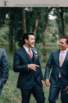 three men in suits are laughing and walking through the woods together with trees behind them