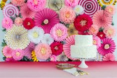 a white cake sitting on top of a table covered in pink and yellow paper flowers