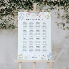 a seating chart on an easer with flowers and greenery behind it, in front of a wall