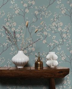 two white vases on a wooden table with flowers in the middle and wallpaper behind them