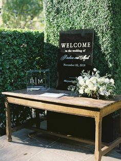 a wooden table with flowers on it and a welcome sign in front of some bushes