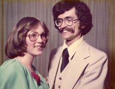 a man in a suit and tie standing next to a woman who is wearing glasses