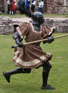 a man dressed in medieval clothing holding a stick and wearing a helmet while running across a field