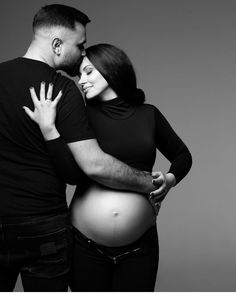 a pregnant couple cuddles and kisses while posing for a black and white photo