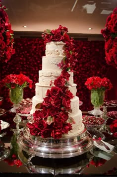 a white wedding cake with red flowers on top is surrounded by silver dishes and glassware