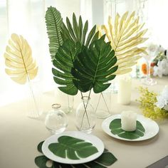 the table is set with white plates and green leaves in glass vases on it