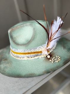 "The perfect sea-foam green hat has been hand painted, and distressed. It's a smaller fedora and has a blue denim paisley band, wrapped in lace and gold velvet. A pearl and gold chain with a pearl tassel is attached. Tied with pink twine is a white feather and brown duck tail feathers.   SIZE: Fedora Hat is OSFA Wired brim measuring 3 3/4\" and a crown height of 4 3/4\". The Fedora Hat has a adjustable band with drawstring which allows for a perfect fit. Materials: This hat is made from a blend of 65% cotton and 35% polyester, providing a comfortable and durable construction. It also includes a faux leather band, suede cord, and with feathers. BENEFITS: - All-Season: This Fedora Hat is suitable for all seasons, allowing you to elevate your style year-round. - Versatile: This wide brim fedo Vintage Fedora Mini Hat For Festivals, Vintage Fedora For Festival, Vintage Fitted Fedora For Festival, Fitted Vintage Fedora For Festival, Vintage Flat Brim Fedora For Festivals, Green Fedora For Kentucky Derby, Handmade Vintage Mini Fedora, Vintage Brimmed Fedora For Festivals, Vintage Summer Felt Hat For Country Events