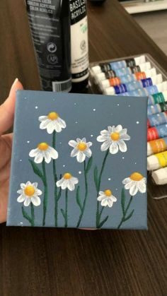 a person is holding up a painted canvas with daisies on it and crayons in the background