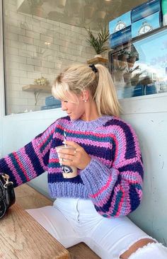 a woman sitting on a bench holding a cup and looking at her cell phone while wearing a purple striped sweater