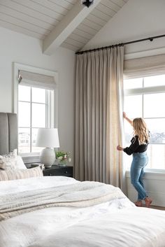 a woman standing in front of a window next to a white bed with beige drapes