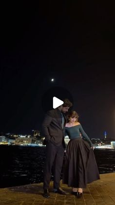 a man and woman standing next to each other in front of the water at night