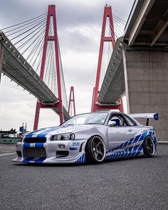 a silver car with blue stripes parked in front of a red and white suspension bridge