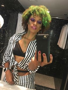 a woman with green hair is looking at her cell phone in the mirror while wearing black and white striped clothing