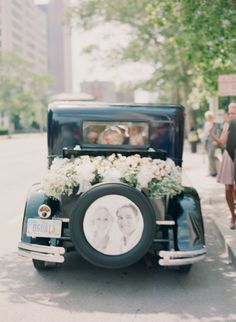 an old fashioned car with flowers on the front is parked near a curb and people are walking down the street