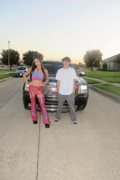 a man and woman standing next to a car in front of a driveway with grass