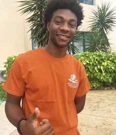 a young man with an orange shirt giving the thumbs up sign in front of some bushes